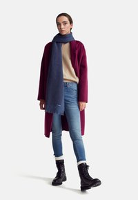 Abrigo burdeos, suéter beige, bufanda azul marino, jeans azul claro y botas negras con cordones y calcetines blancos; conjunto casual con texturas variadas.