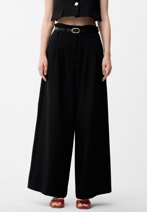 Femme portant un pantalon noir taille haute à jambes larges avec une ceinture noire et un top court noir, assorti à des sandales rouges à bout ouvert.