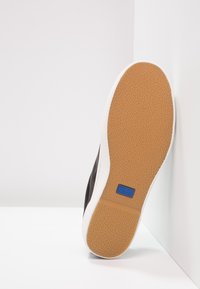 Černé tenisky s texturovanou hnědou gumovou podrážkou a bílou mezipodešví. Na boku se nachází logo Keds v modré barvě.