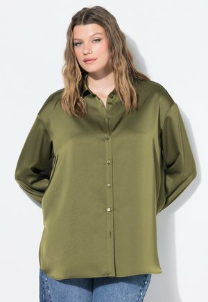 Junge Frau mit welligem Haar, die ein olivgrün-satiniert Button-Up-Hemd und eine blaue Jeans trägt, steht vor einem schlichten weißen Hintergrund.