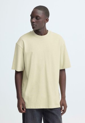 Beige T-Shirt mit kurzen Ärmeln, Rundhalsausschnitt, weiter Passform und strukturierter Stoff. Kombiniert mit dunkelgrauen Hosen, Modell ist leicht abgewandt.