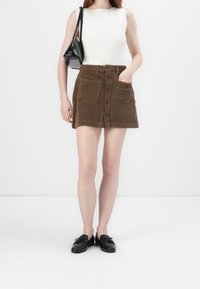 Femme portant un haut blanc sans manches, une jupe en velours côtelé marron à boutons sur le devant, des mocassins noirs, et portant un sac à main noir à l'épaule.