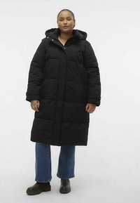 Cappotto imbottito nero con cappuccio, dotato di una zip frontale, tasche laterali e lunghezza fino al ginocchio. Indossato con jeans blu e stivali neri.