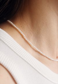 Pērļu kaklarota ar mazām, vienveidīgām baltām pērļu kuloniņām, kas maigi atpūšas uz kakla, pret vieglu ādas toni. Redzams teksturēts balts audums.