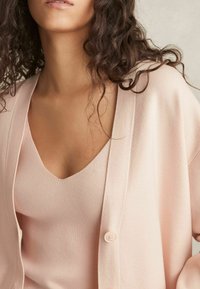 Cardigan rosa chiaro sopra un top a scollo a V coordinato, caratterizzato da una texture morbida e una chiusura a bottoni sul cardigan. Design semplice, senza motivi.