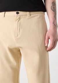 Pantalons en coton beige avec une texture lisse, poches standard et fermeture à bouton à l'avant, présentés avec une chemise noire.