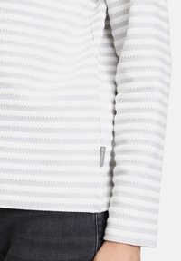 Haut blanc à manches longues avec des rayures grises horizontales, tissu texturé et une petite étiquette sur le côté. Porté avec un denim foncé.