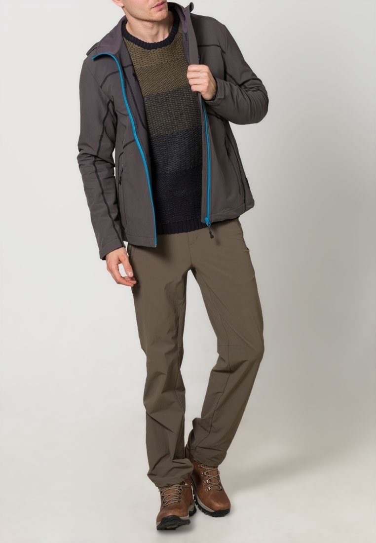 Homme portant une veste marron avec des détails de fermeture éclair bleus, un pull color-block, un pantalon marron et des chaussures de randonnée marron, debout devant un fond uni.