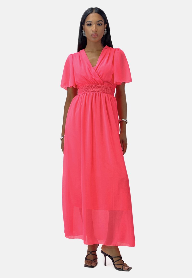 Robe maxi en chiffon rose vif, avec de courtes manches flottantes, une taille smockée et une silhouette fluide. Portée avec des sandales noires à brides.