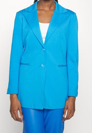 Kobieta ubrana w jasno niebieską marynarkę z dwoma guzikami, w połączeniu z dopasowanymi niebieskimi spodniami i białą bluzką pod spodem.