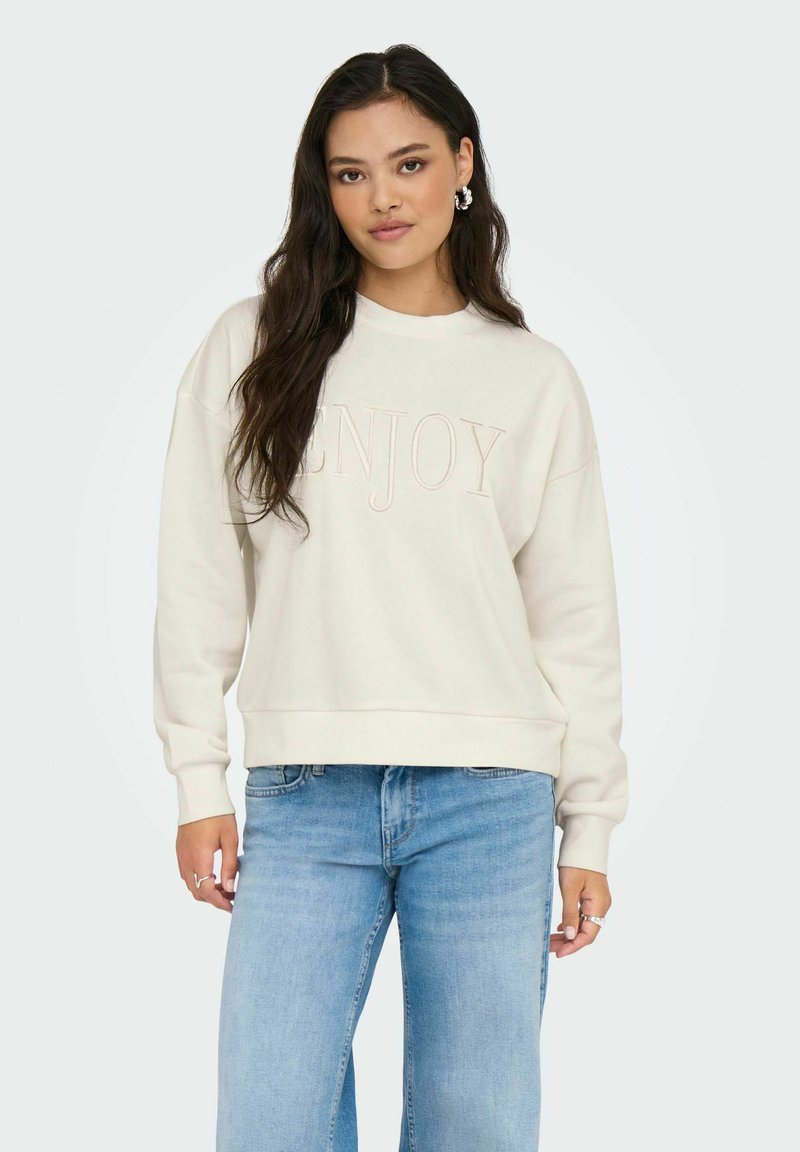 Femme aux longs cheveux noirs portant un sweat-shirt blanc avec le texte "ENJOY" et un jean bleu clair, se tenant devant un fond clair uni.