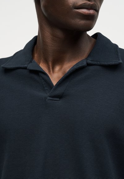 Gros plan d'une personne portant un polo texturé bleu marine foncé avec un col ouvert, montrant le cou et le bas du visage.