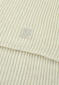 Tissu tricoté couleur crème avec une texture côtelée et une sensation douce, arborant une petite étiquette carrée avec un motif de fleur brodé.