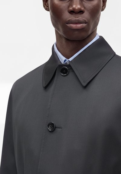 Gros plan d’une personne portant un manteau boutonné gris foncé sur une chemise bleu clair à col, sur un fond uni.