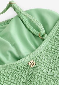 Tessuto verde con trama texturizzata, una fodera interna liscia, spalline in pizzo e un accento in metallo dorato all'escollatura.
