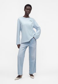 Femme debout, main dans la poche, portant une chemise bleu clair à manches longues, un jean large bleu clair avec des ourlets effilochés et des chaussures pointues beiges.