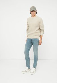 Pulover tricotat bej, blugi slim bleu deschis, șapcă gri și adidași albi cu accente bej. Design simplu, texturat, fără modele.
