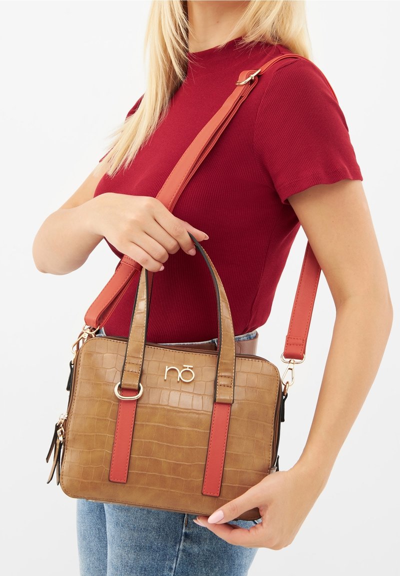 Femme portant un haut rouge à manches courtes et un jean bleu tient un sac à main marron à texture crocodile avec des lanières rouges sur l'épaule et par les poignées.