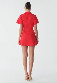 Vrouw in een korte, met een riem gezepte rode jurk, staat met haar rug naar de camera, met een eenvoudige achtergrond die de outfit en haar benen in huidskleurige hakken benadrukt.