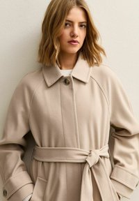 Beżowy płaszcz z paskiem, kołnierzem, raglanowymi rękawami i zapięciem na guziki. Miękka, fakturowana tkanina o gładkim wykończeniu oraz boczne kieszenie.