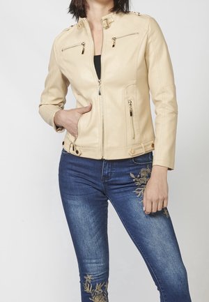 Kvinde iført beige læderjakke med lynlås foran samt lynlåslommer på bryst og sider, kombineret med blå broderede skinny jeans.