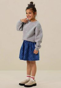 Sweatshirt gris avec broderie florale, associé à une jupe bleue avec un motif de feuilles. Pour compléter le look, des chaussures crème et des chaussettes rayées rouges.