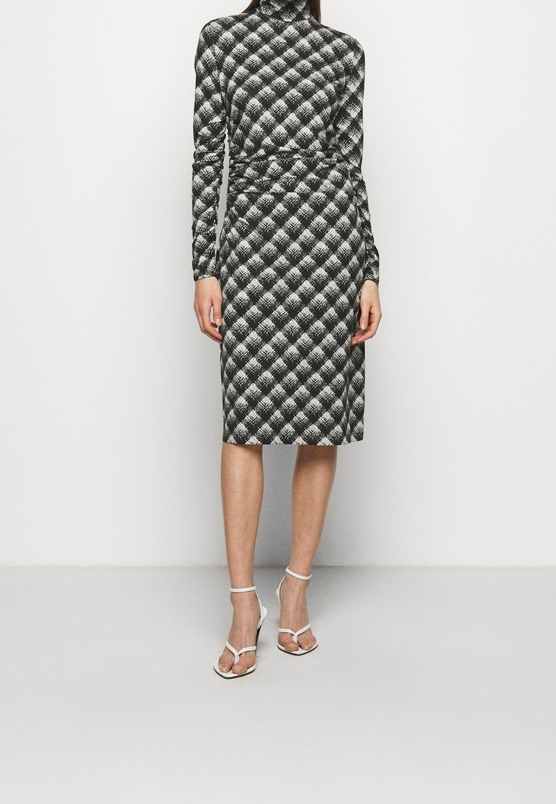 Czarna i biała sukienka w wzory z długimi rękawami, golfem, dopasowaną sylwetką i długością do kolan. Posiada fakturowany wzór w romby.