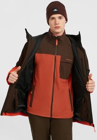 Fleece-gevoerde zip-up jas in bruin en oranje met een hoge kraag, zijzakken en een gebrandmerkt patch. Gecombineerd met een bijpassende bruine beanie.