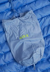 Sacca azzurro chiaro con una texture increspata, caratterizzata dal testo "JJXX" in verde brillante. Posizionata su uno sfondo blu trapuntato.