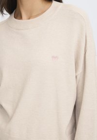 Bež pulover z zaobljenim izrezom in rebrastimi manšetami. Na levi strani prsnega koša ima majhen rožnato vezen srček. Mehka tekstura.