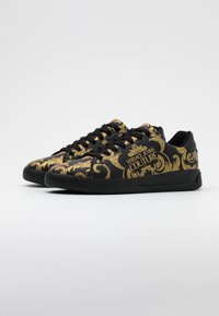 Black sneakers featuring a gold baroque pattern, with black laces and a textured rubber sole. Branding visible on the side.