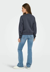 Donna in piedi, leggermente di lato, con felpa blu navy, jeans azzurri chiari, sneakers bianche e orecchini a cerchio argentati su uno sfondo neutro.