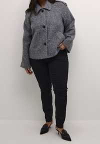 Grå uldblandet jakke med store knapper, spids krave og to sidelommer; kombineret med sorte, tætsiddende bukser og spidse hæle.