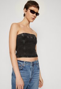 Top senza spalline in denim nero con design strutturato e dettaglio del logo rosso; abbinato a jeans azzurri a vita alta. Texture liscia.