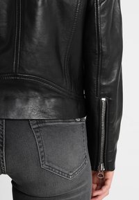 Sort læderjakke med sølvfarvet lynlås på ærmet iført over grå denimjeans med synlige baglommer.