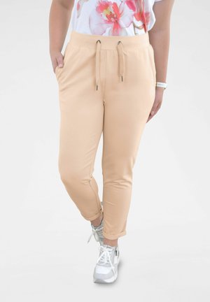 Persoon draagt beige broek met trekkoord, witte en grijze sneakers en een mouwloze top met bloemenprint, staat met één hand in de zak.