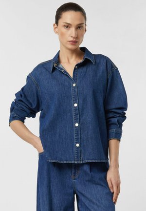 Kobieta ubrana w niebieską dżinsową koszulę z długim rękawem i srebrnymi guzikami oraz pasujące niebieskie dżinsowe spodnie, stojąca z jedną ręką w kieszeni.