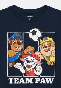 Tengerészeti kék póló, amelyen ruhás rajzfilm kutyák láthatók focilabdával, alatta a "TEAM PAW" felirat. Rövid ujjú és kerek nyakú kialakítás.