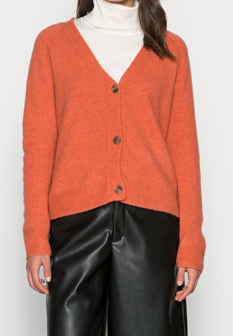 Orange stickad cardigan med V-ringning, fem bruna knappar, ribbade ärmslut och nederkant, bärs över en vit polotröja och matchas med svarta läderbyxor.