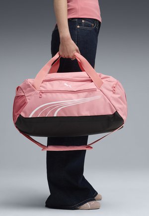 Persona sosteniendo una bolsa de deporte Puma rosa y negra con asas dobles, usando jeans oscuros y zapatos beige con el logo de Puma.