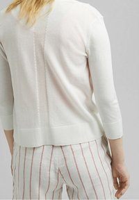 Femme portant un pull en maille blanc avec une bande en maille verticale dans le dos et un pantalon blanc avec des rayures verticales roses.