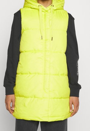 Osoba ubrana w neonowo żółtą pikowaną kamizelkę bez rękawów z kapturem, noszoną na czarnej koszuli z długim rękawem, stojąca na jednolitym tle.