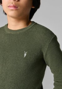 Maglione a coste verde scuro con scollo rotondo e un piccolo emblema bianco con la testa di una capra sul lato sinistro del petto.