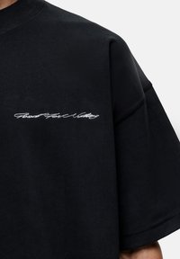 Black oversized t-shirt with short sleeves, featuring white embroidered text on the left side. Made from soft cotton fabric.
