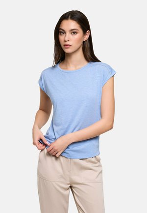 Mujer de cabello largo y castaño lleva una camiseta de manga corta azul claro y pantalones beige, ajustando el dobladillo de la camiseta con ambas manos.