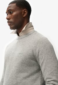 Hellgrauer Pullover mit geripptem Halsausschnitt und Raglanärmeln, über einem beigen Hemd mit Kragen getragen. Weiche Textur, minimale Markenkennzeichnung sichtbar.