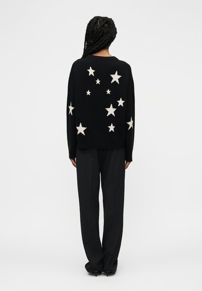 Persona con capelli intrecciati vista di spalle, che indossa un maglione nero decorato con stelle bianche di varie dimensioni e pantaloni neri.