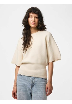 Kvinde med langt krøllet mørkt hår iført en løs cremefarvet kortærmet sweater og lyseblå højtaljede jeans mod en ensfarvet baggrund.