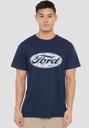 Man wearing navy blue T-shirt with white oval Ford logo, standing against a plain light gray background.