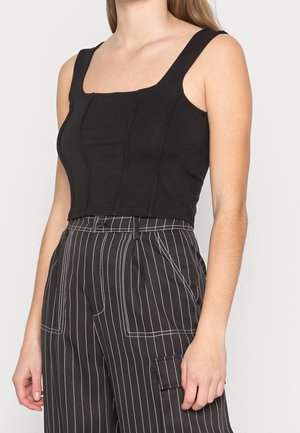 Vrouw in een aansluitend zwart mouwloos crop top en een hoge taille zwarte broek met witte verticale streepjes en opgestikte zakken.
